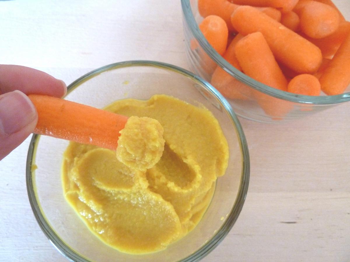 Hand dipping a baby carrot into a homemade Nutritarian turmeric hummus.