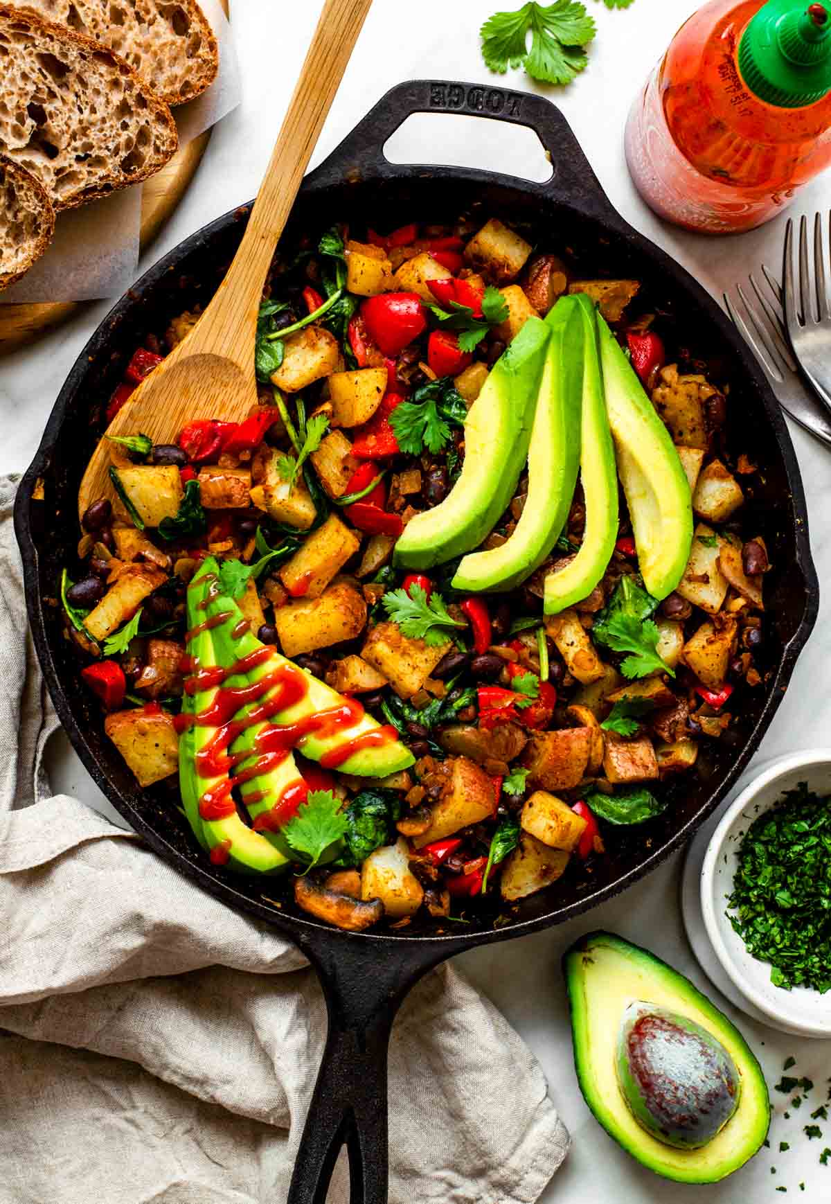 Vegetable hash in a cast iron skillet, topped with avocado slices, fresh cilantro leaves, and a drizzle of hot sauce.