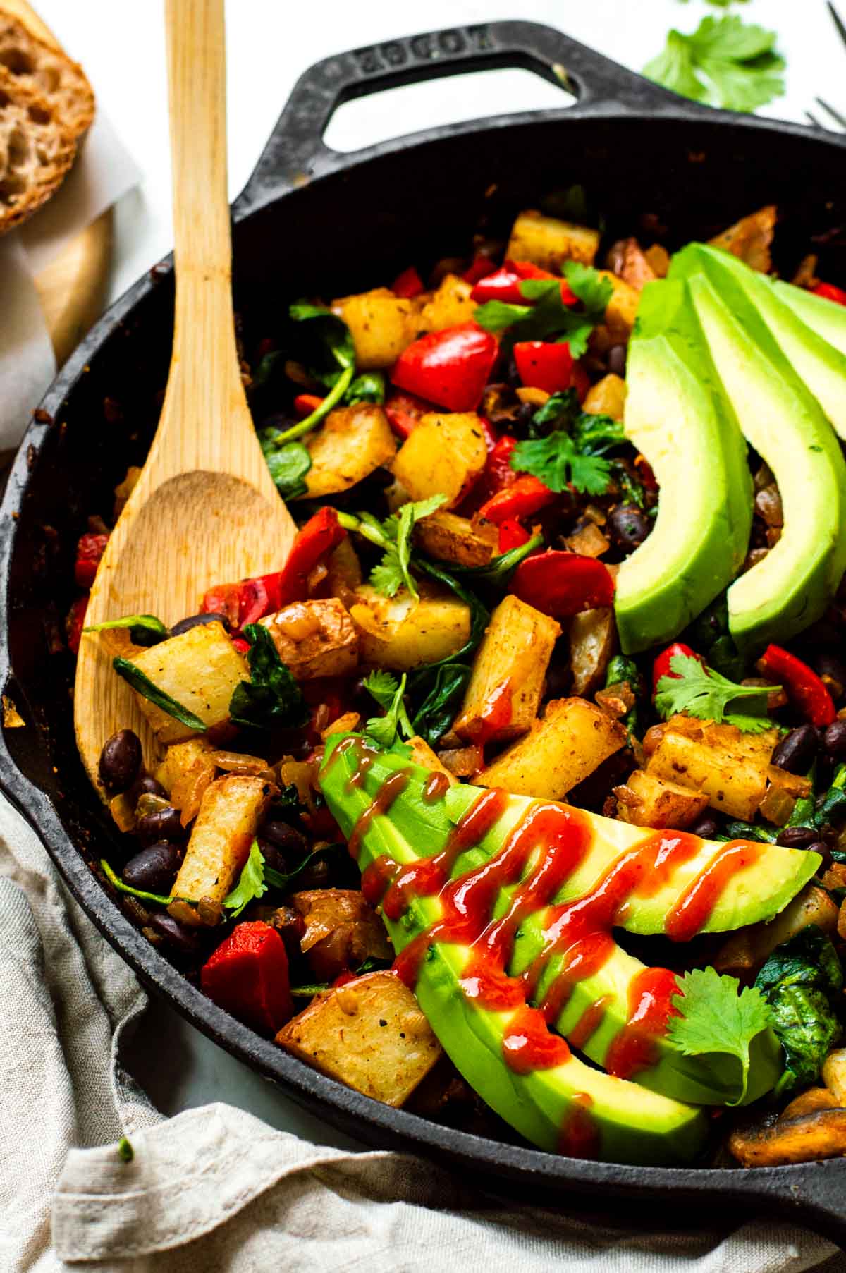 Vegetable hash in a cast iron skillet, topped with avocado slices, fresh cilantro leaves, and a drizzle of hot sauce.