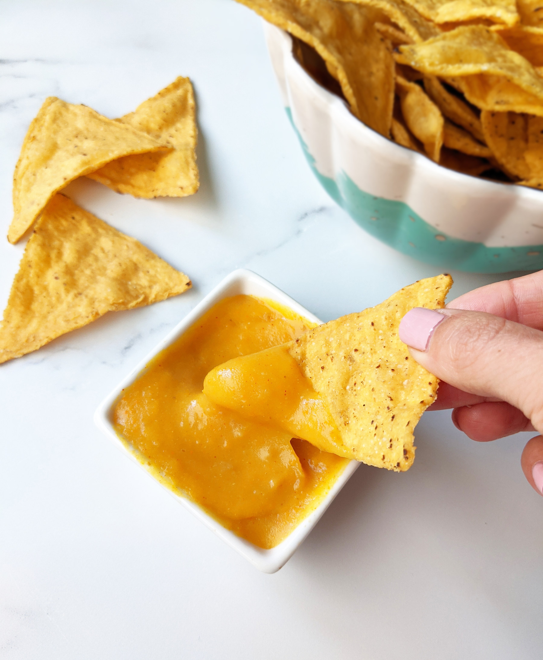 A hand dipping a tortilla chip into a small serving dish filled with vegan nacho cheese sauce.