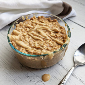 Glass bowl filled with overnight oats and drizzled with peanut butter and a spoon beside it.