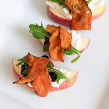 Apple slices covered with vegan cream cheese, a drizzle of balsamic glaze, an arugula leaf, and wrapped in carrot bacon