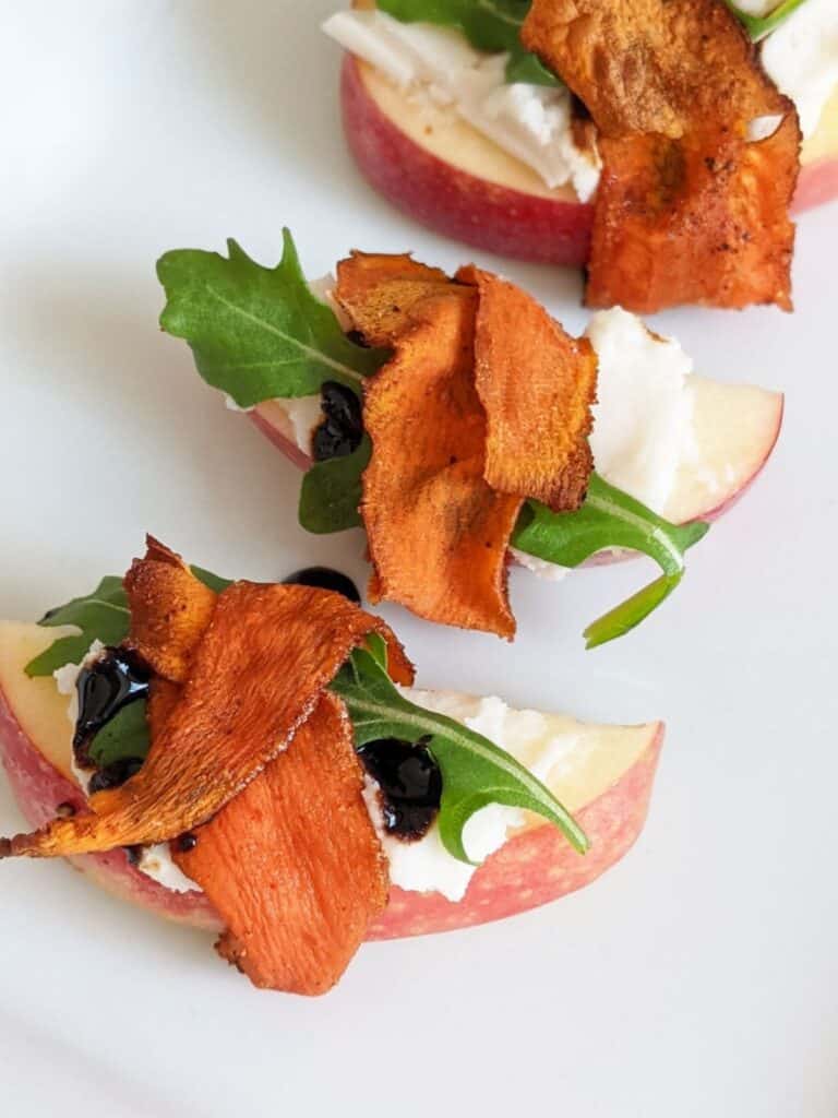 Apple slices covered with vegan cream cheese, a drizzle of balsamic glaze, an arugula leaf, and wrapped in carrot bacon