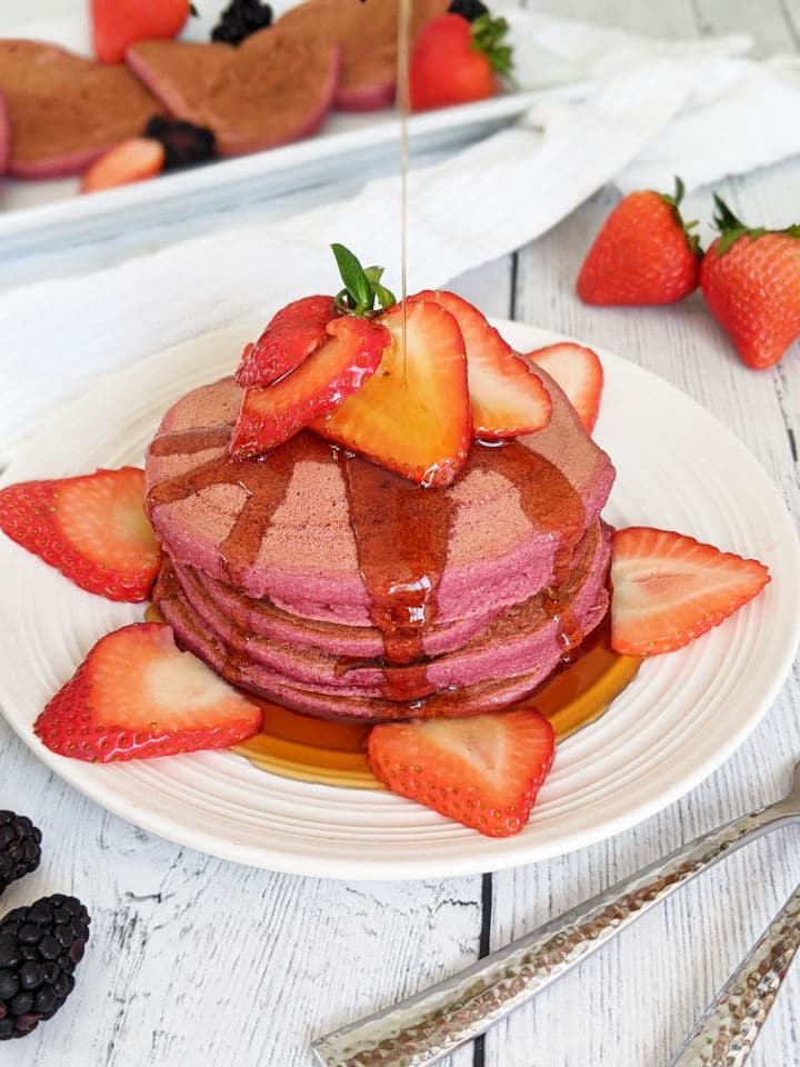 A stack of vegan pancakes pink from beets and drizzled with maple syrup, topped with strawberries