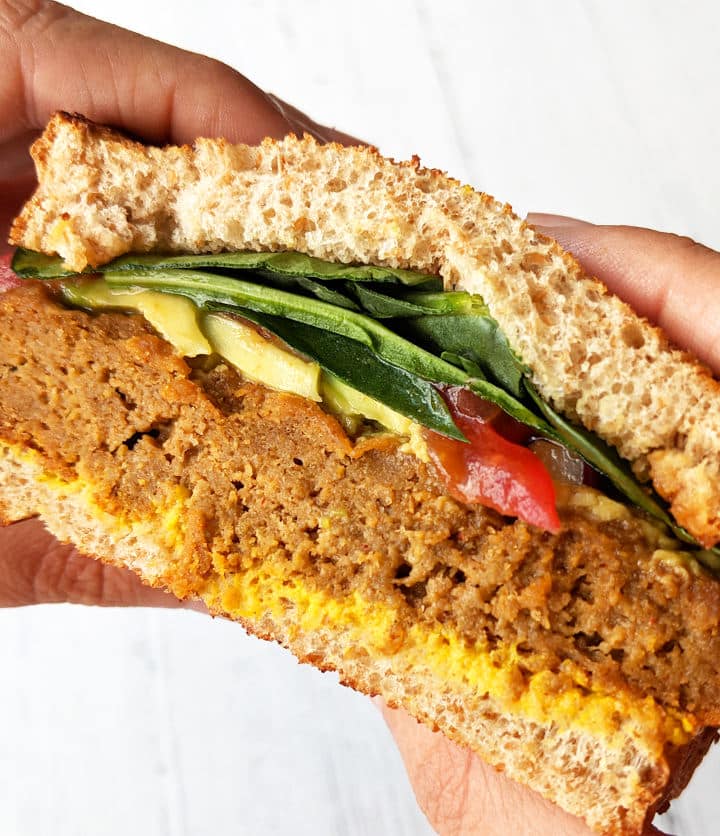 Up close shot of hands holding a BBQ Seitan Sandwich topped with veggies, avocado, and mustard