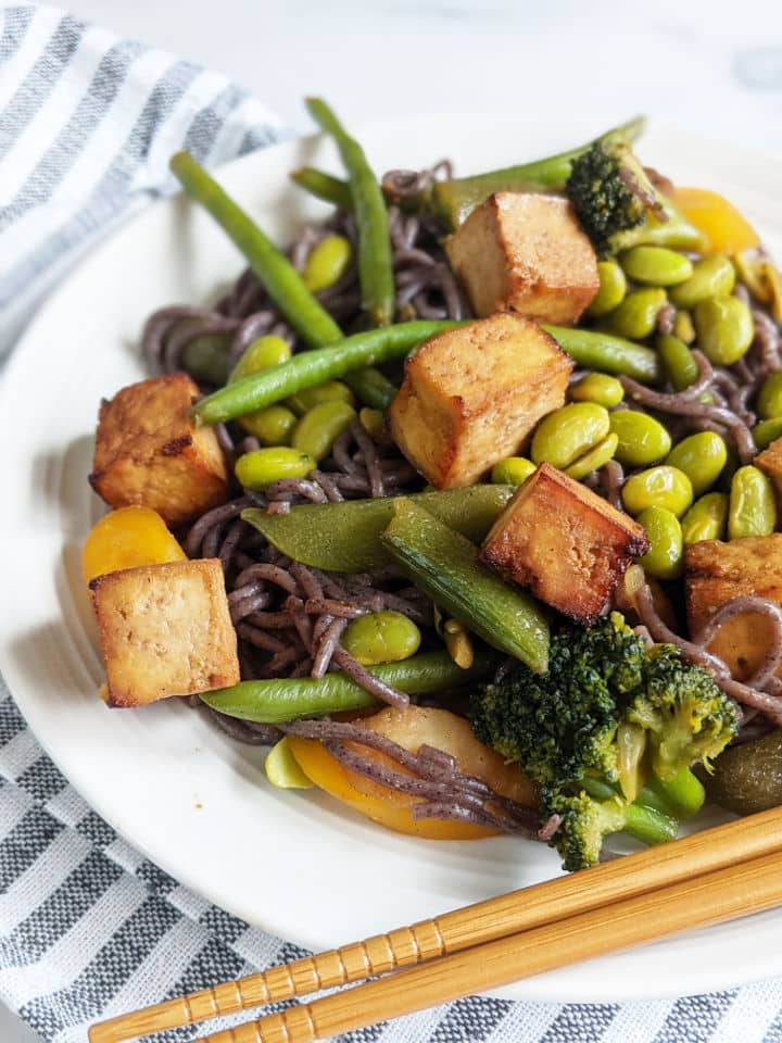 Tofu and vegetable stir fry with black rice noodles on a plate with chopsticks