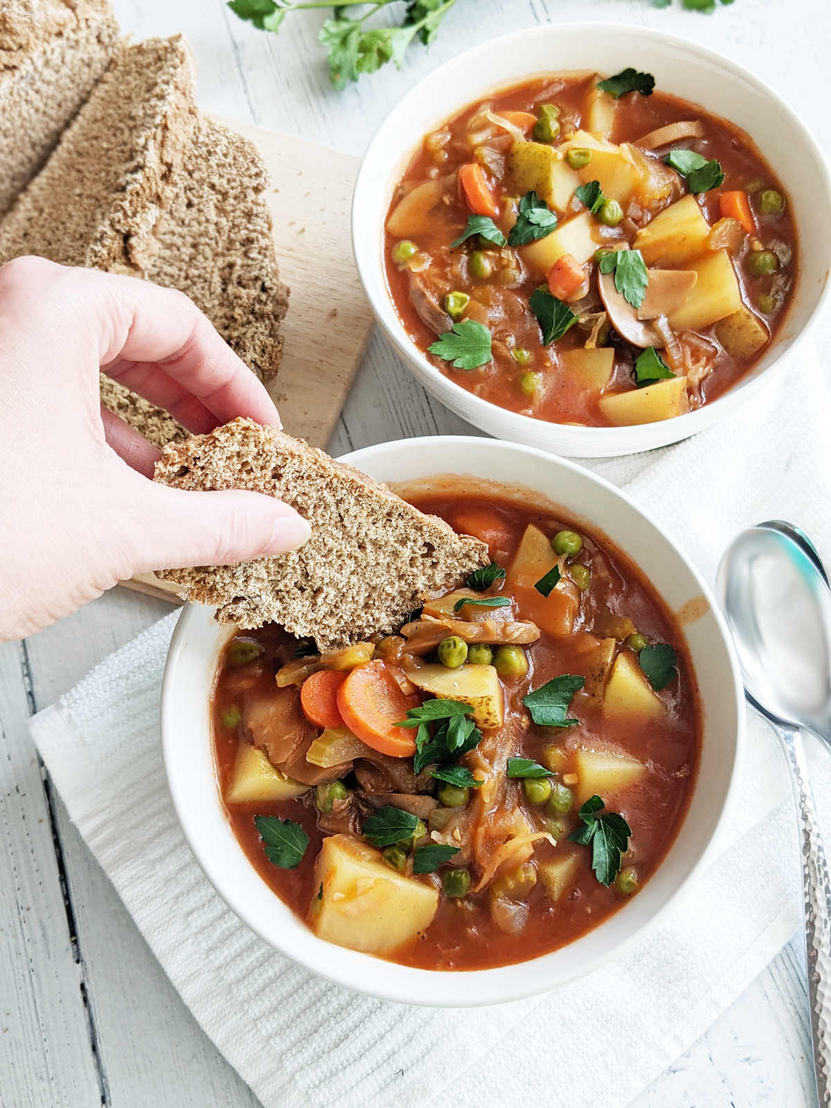 A bowl of vegetable stew topped with parsley with a hand dipping a slice of soda bread into the stew.