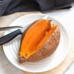A sliced open cooked sweet potato on a plate with a fork and knife.