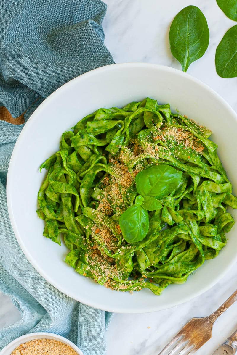 Bowl with fettuccine pasta covered in green sauces sprinkled with yellow flakes and a pair of basil leaves.
