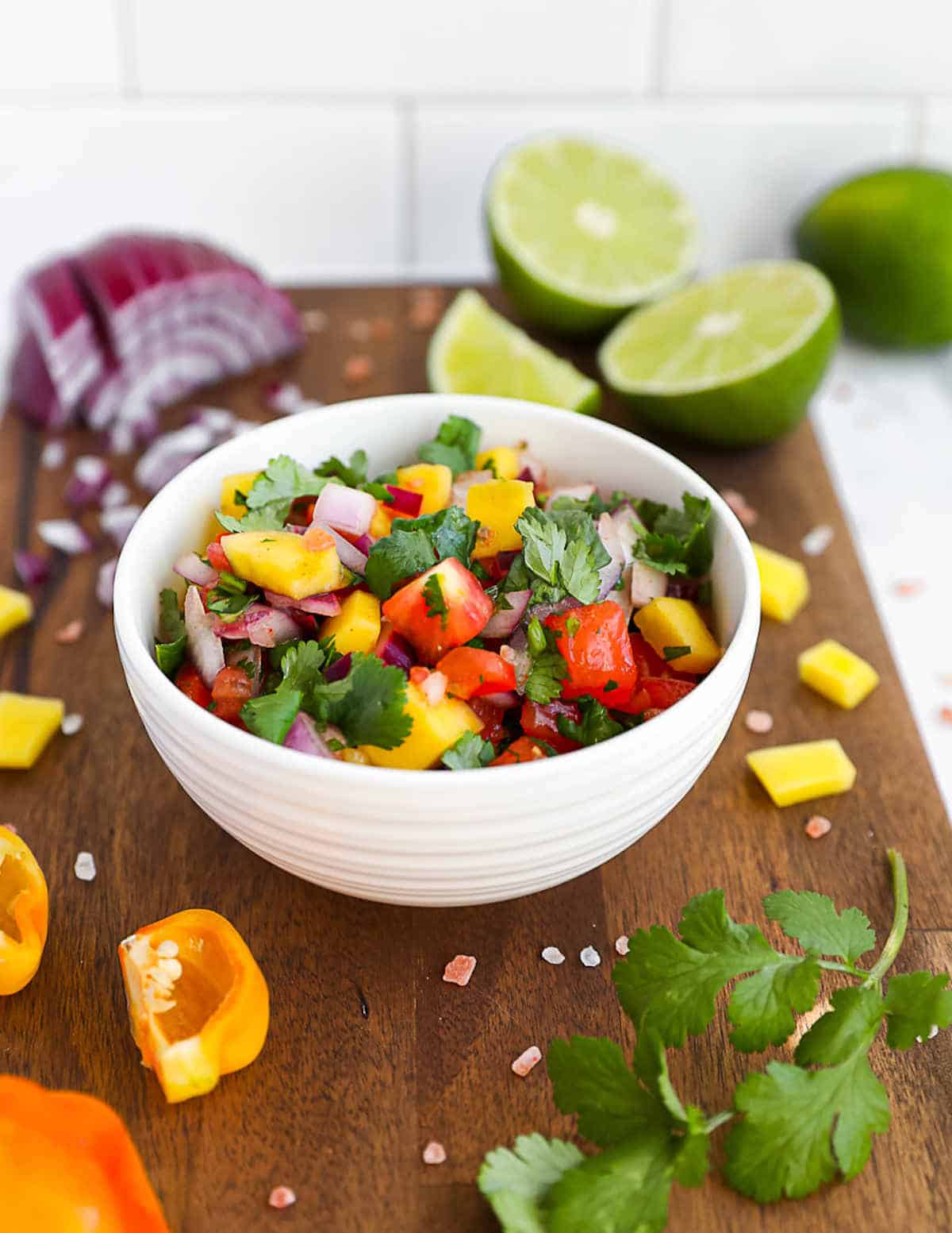 Mango salsa in small bowl on a cutting board surrounded by chopped ingredients.