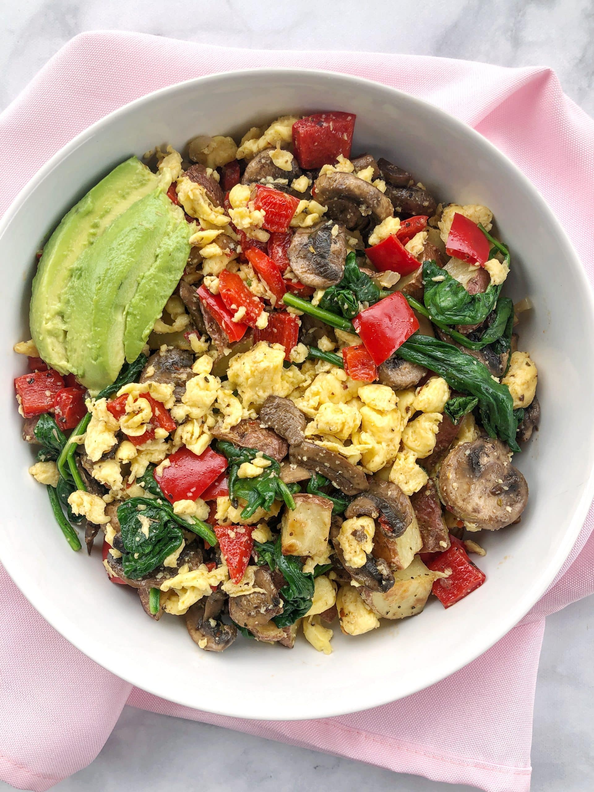 White bowl on pink napkin filled with vegan hash, veggies and avocado.