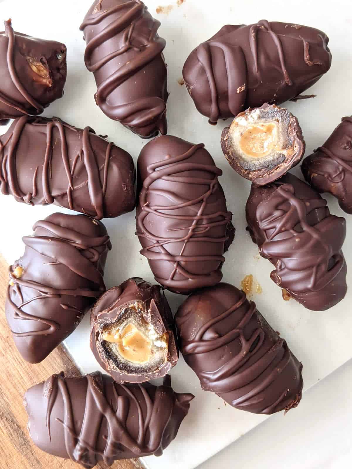 Dates stuffed with peanut butter and covered with chocolate sitting on a serving board with a couple dates cut in half.