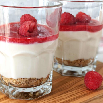 Two no bake vegan cheesecakes in glasses topped with raspberry sauce and fresh raspberries.
