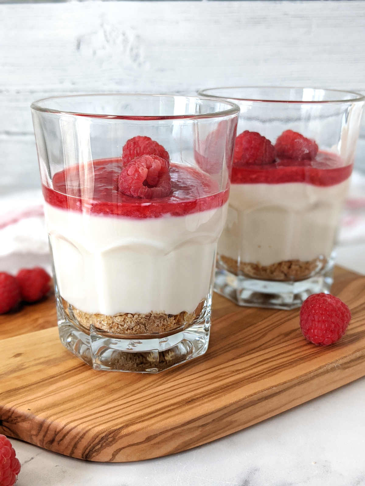 Two no bake vegan cheesecakes in glasses topped with raspberry sauce and fresh raspberries.