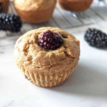 Vegan Blackberry Muffin sitting on light surface with fresh blackberries next to it and a cooling rack holding more muffins behind it.
