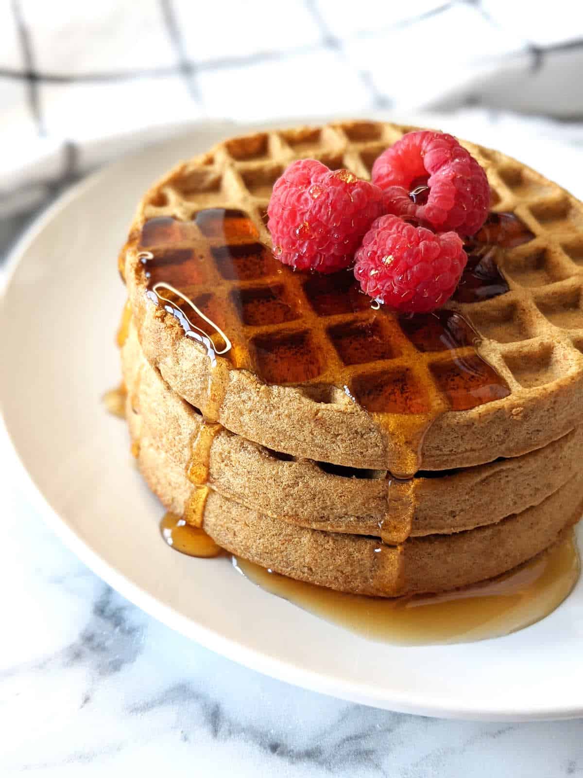 Air fried frozen waffles stacked on a plate topped with raspberries and maple syrup.