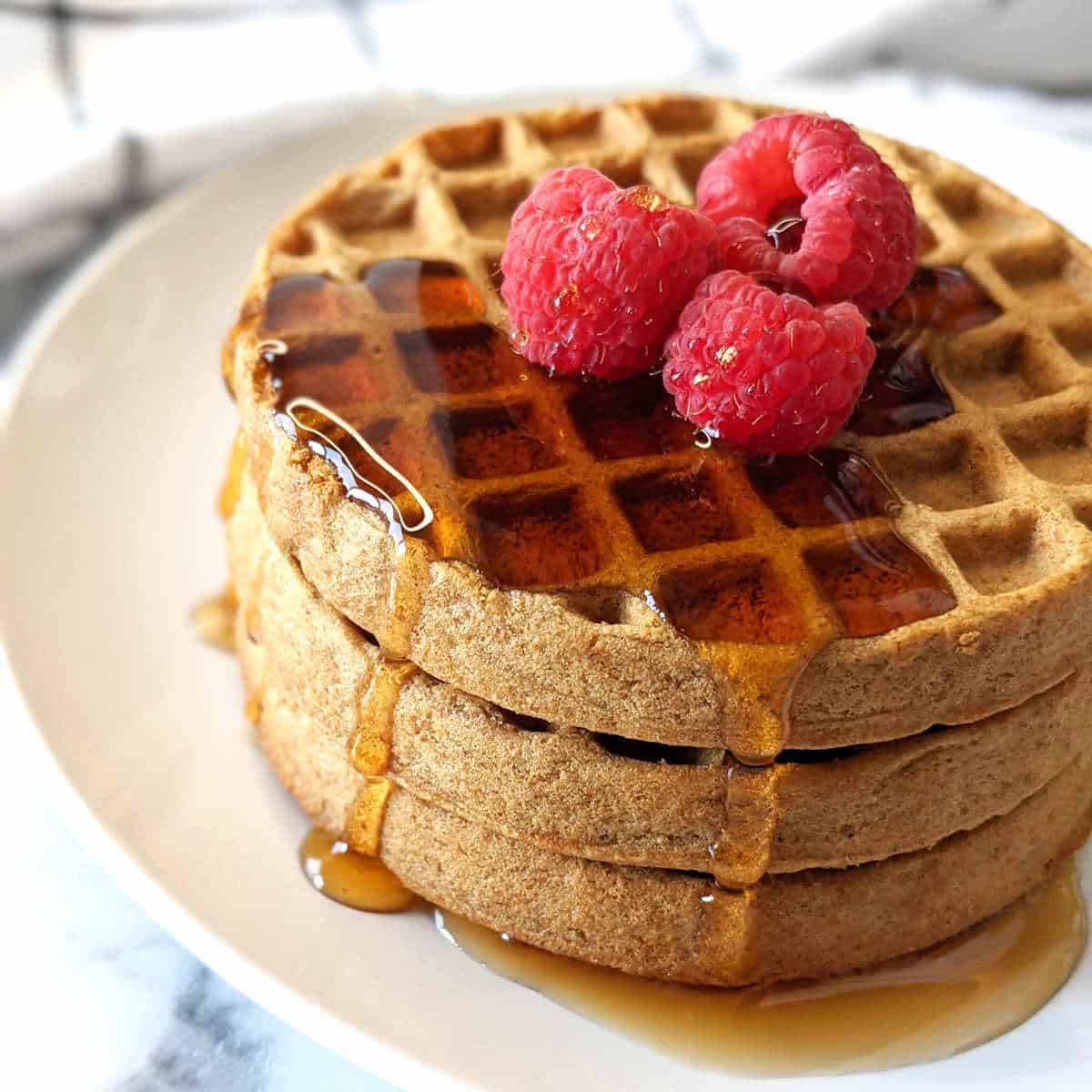 Air fried frozen waffles stacked on a plate topped with raspberries and maple syrup.