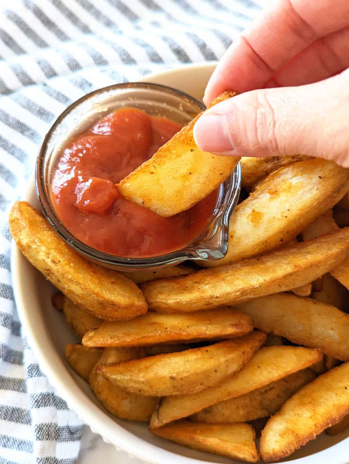 Hand dipping a potato wedge into a small bowl of ketchup.