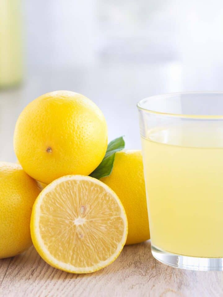 Lemons stacked in a pile with one sliced in half next to a glass of lemon juice.