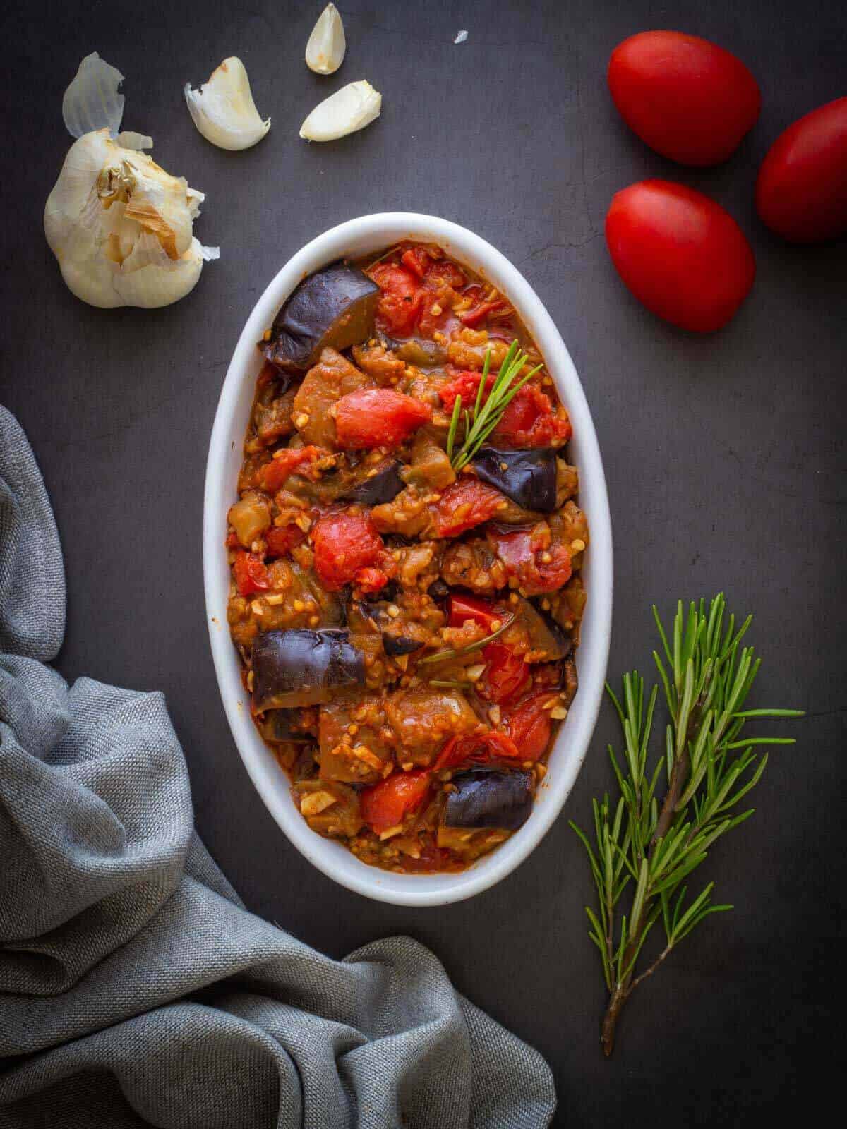 Eggplant stew in a serving dish surround by ingredients and a cloth napkin.