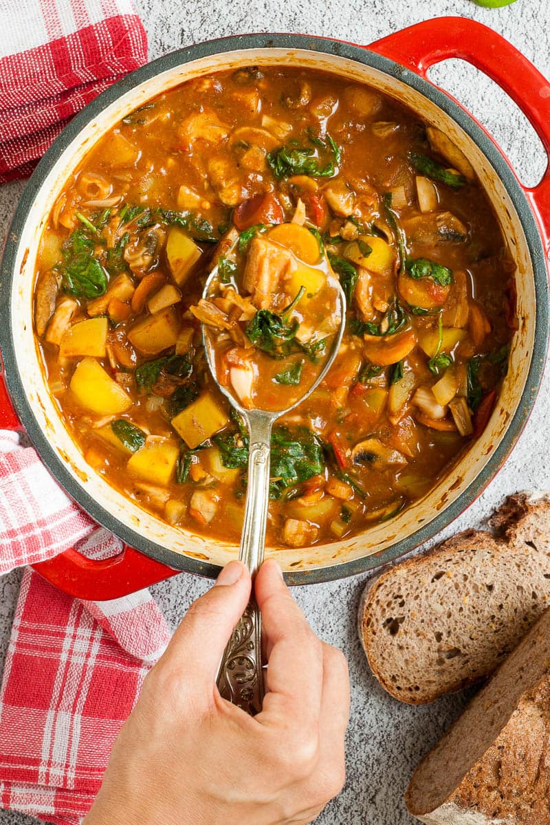 Red Dutch oven from above with a stew where you can see chopped potatoes, spinach leaves, carrot slices, tomatoes, and mushroom slices. Slices of bread is next to it. A hand is holding a spoon taking some from the middle.