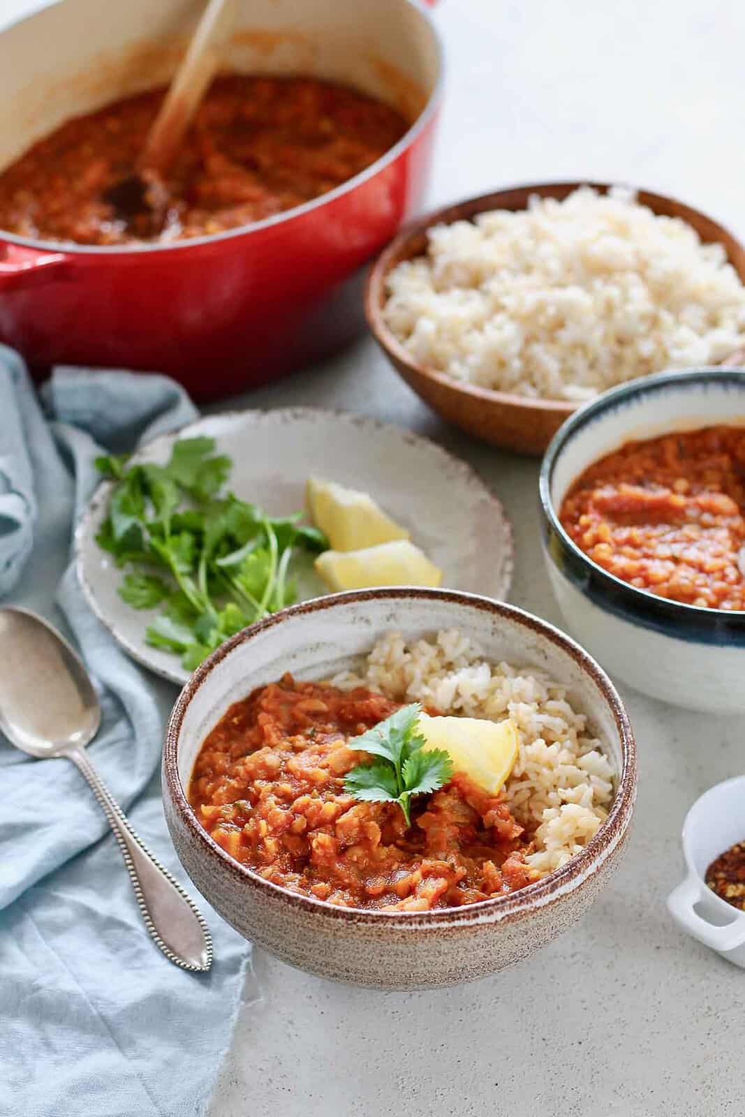Red lentil stew in a bowl with rice surround by dishes filled with rice, stew, and garnishes.