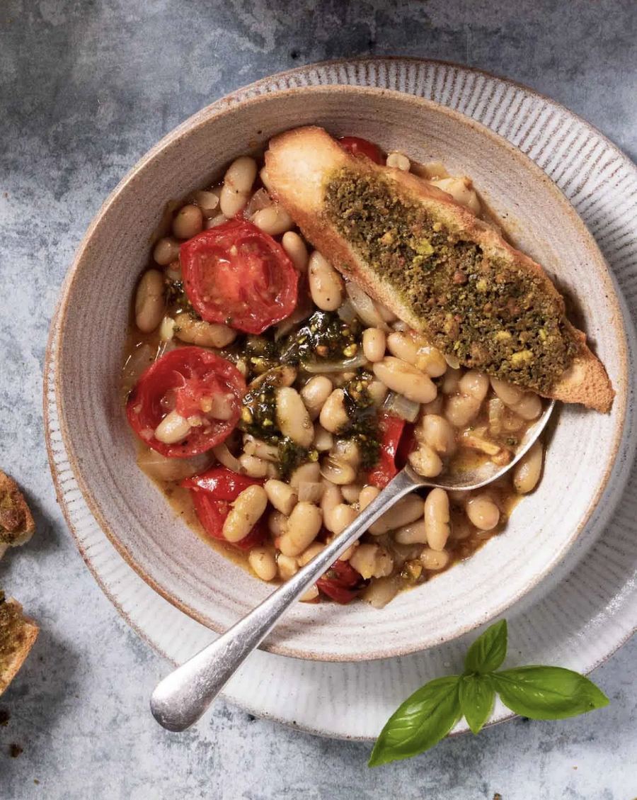 Bowl of white bean stew with roasted tomatoes, served with a spoon and a slice of bread.