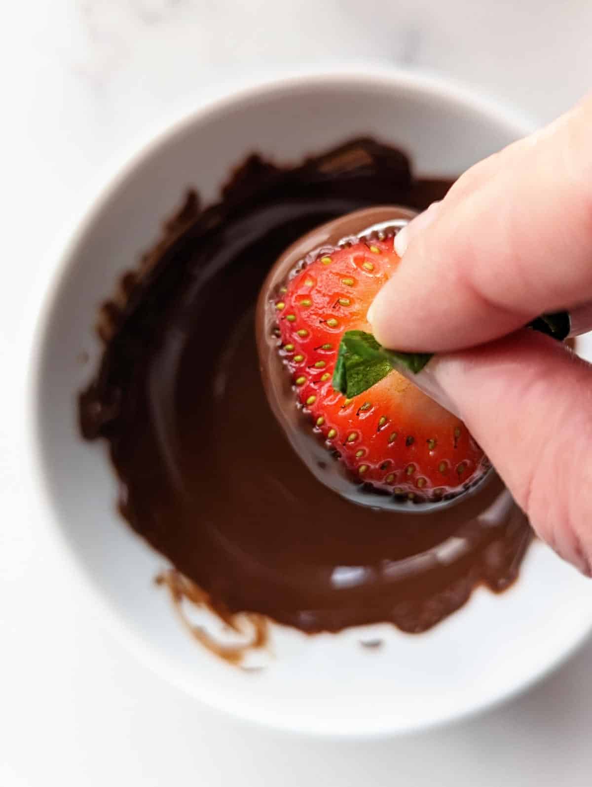 Hand holding dipped strawberry over chocolate bowl to allow excess chocolate to fall off.