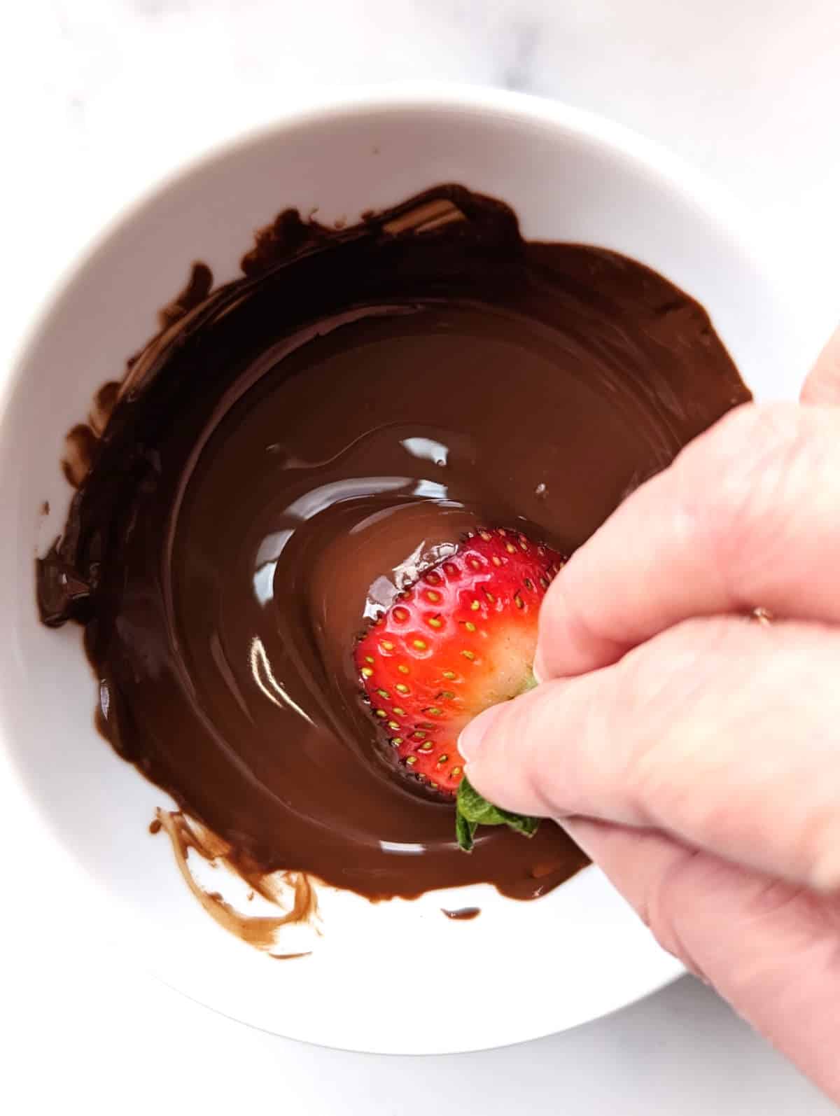 Hand dipping a strawberry into melted chocolate.