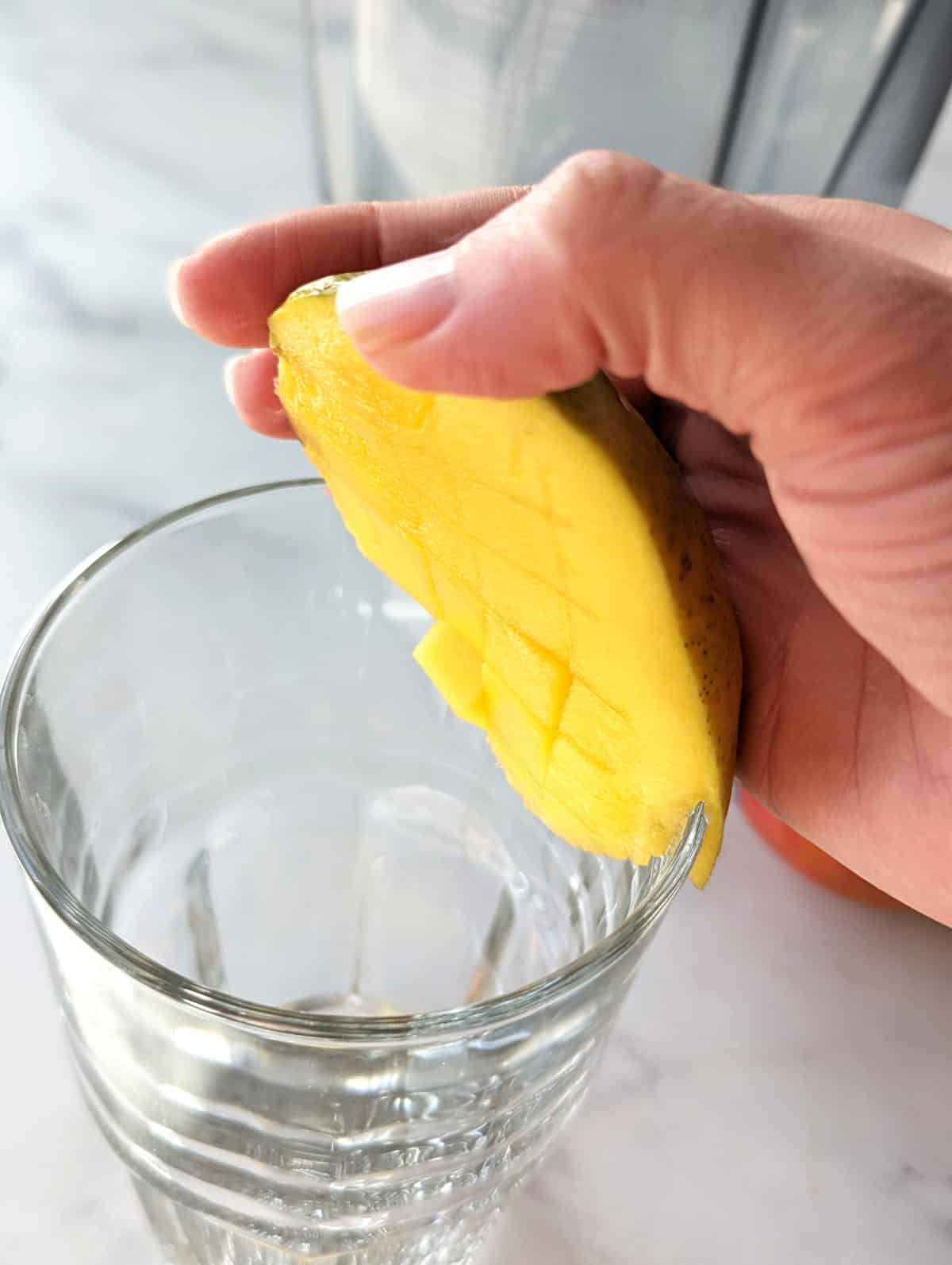 Hand holding a slice of mango against a glass in the process of separating the mango flesh from the peel.