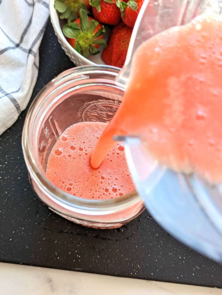 A mason jar with a blender container pouring the strawberry juice into it.