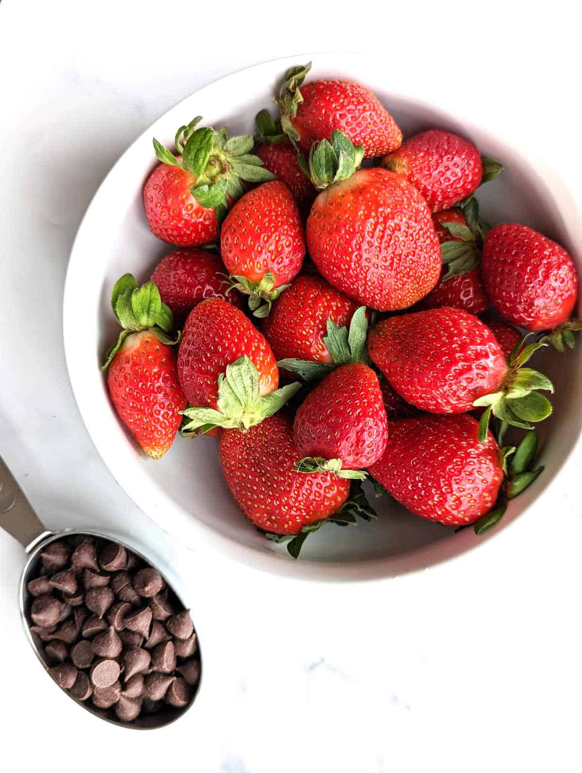 Fresh strawberries in a bowl beside a measuring cup full of dairy-free chocolate chips.