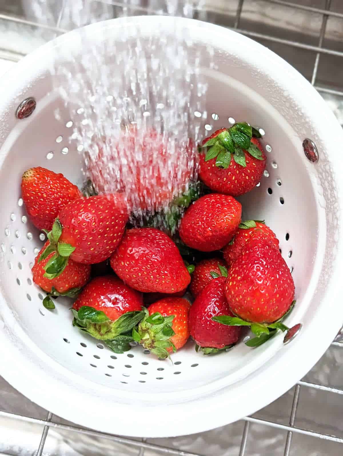 Fresh strawberries in a colander with water spraying down on them.