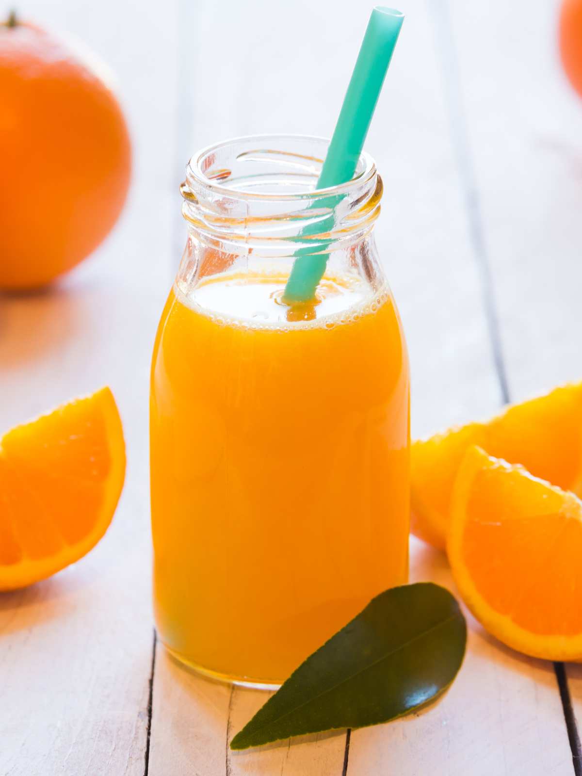 A bottle of freshly squeezed orange juice with a straw next to slices of oranges and an orange leaf.