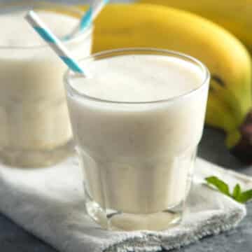 Two glasses of banana milk with straws and bananas in the background.