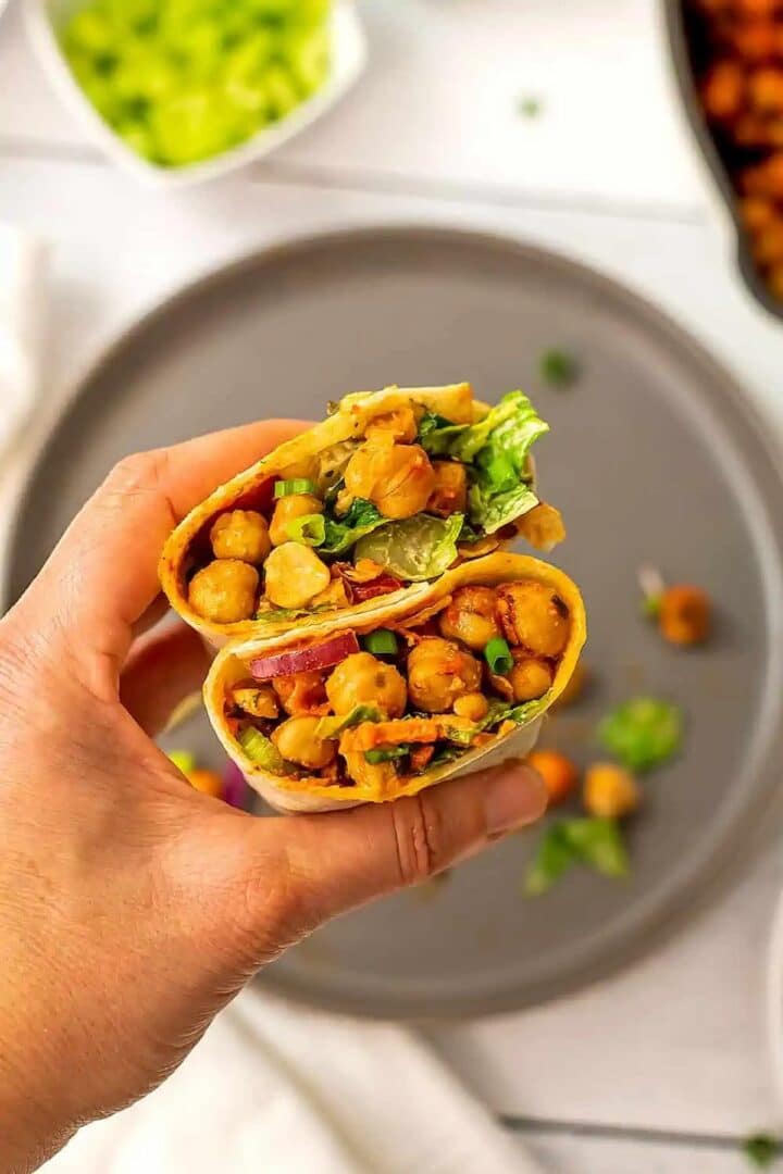 A hand holding up two halves of a buffalo chickpea wrap over a plate.