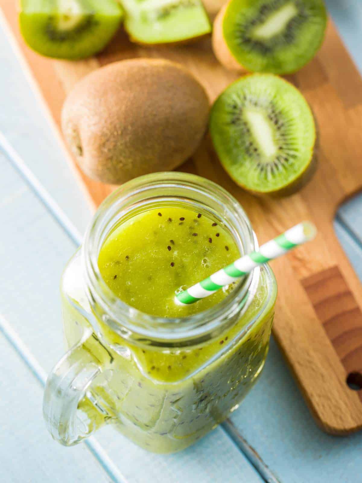 Mason jar filled with kiwi juice, served with a straw, next to several ripe kiwis.