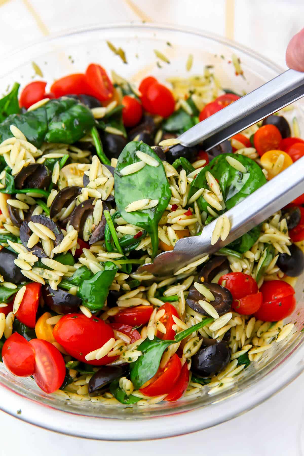 Bowl of pesto orzo salad with veggies and a pair of tongs tossing it all around.