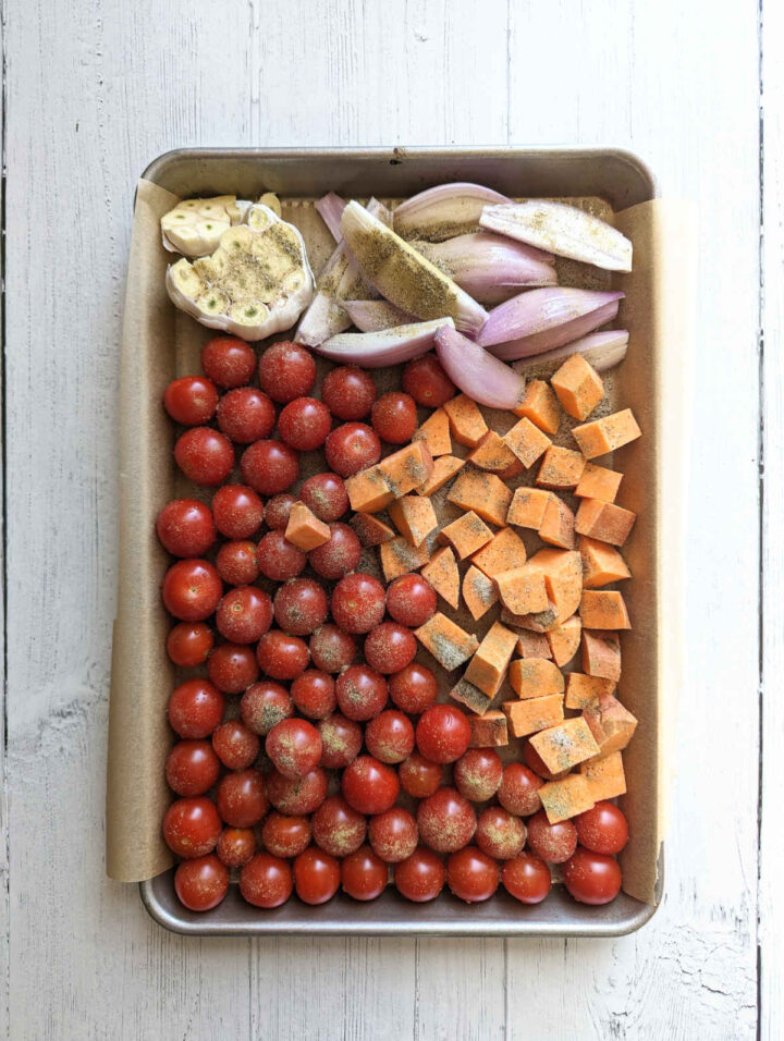 Cherry tomatoes, chopped sweet potato, chopped shallots and a head of garlic spread out on a sheet pan.