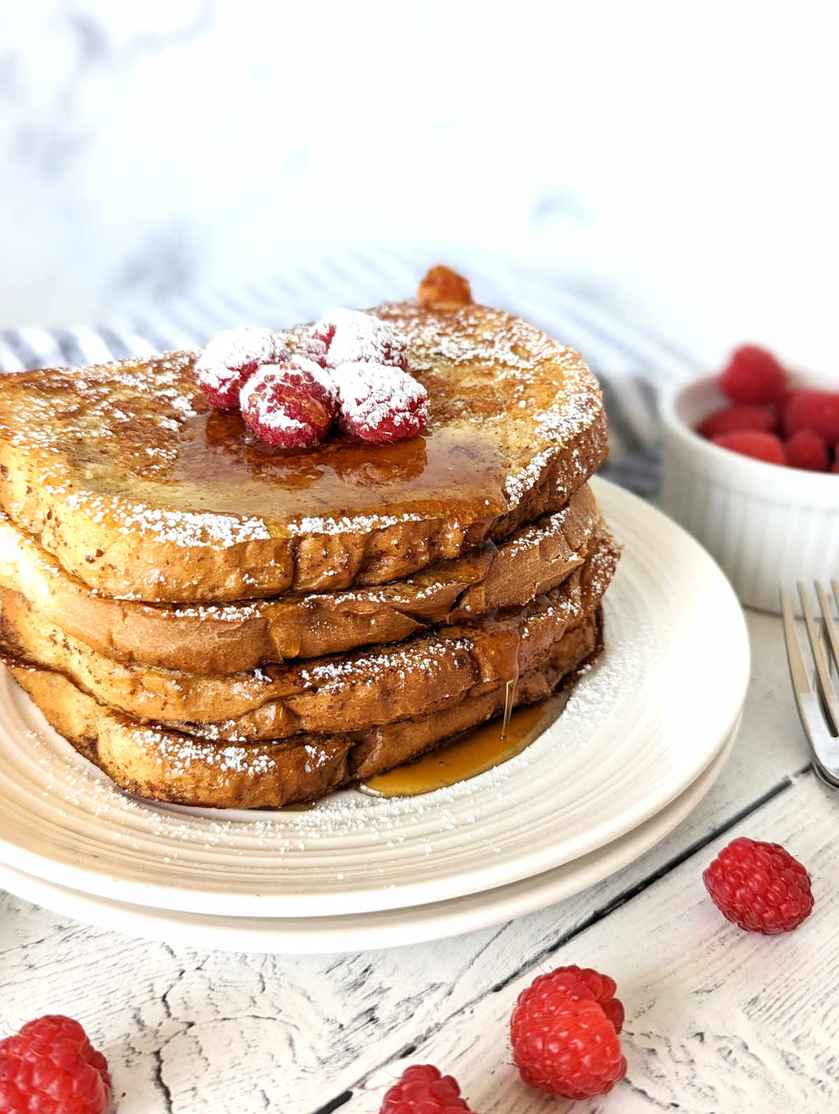 Just Egg French Toast slices stacked on a plate, topped with raspberries, maple syrup and powdered sugar.