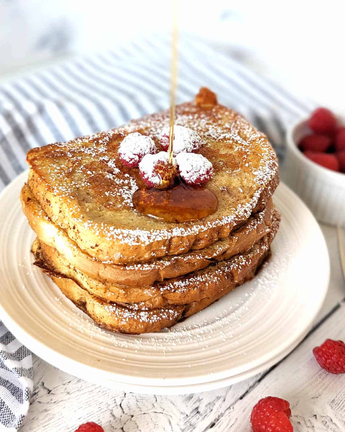 Just Egg French Toast slices stacked on a plate, topped with raspberries and maple syrup in the process of being poured on top.