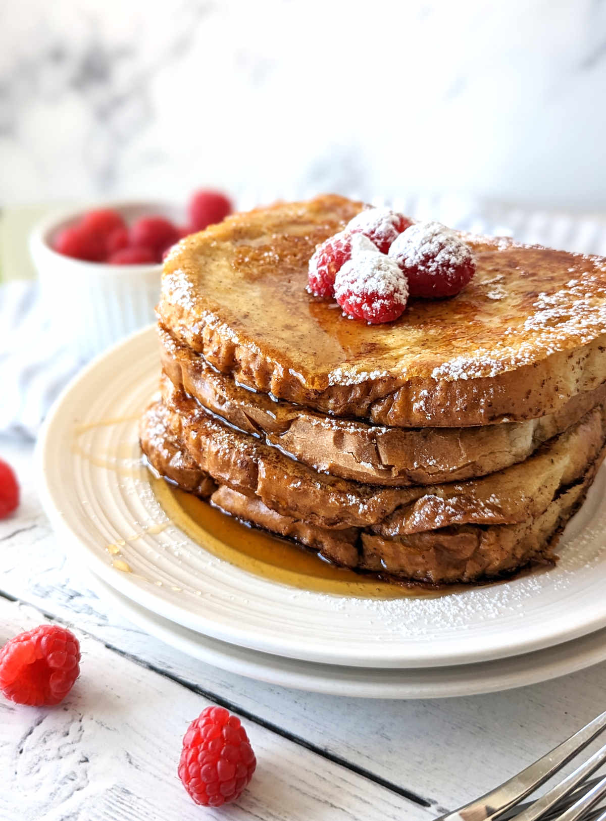Just Egg French Toast slices stacked on a plate, topped with raspberries, maple syrup and powdered sugar.