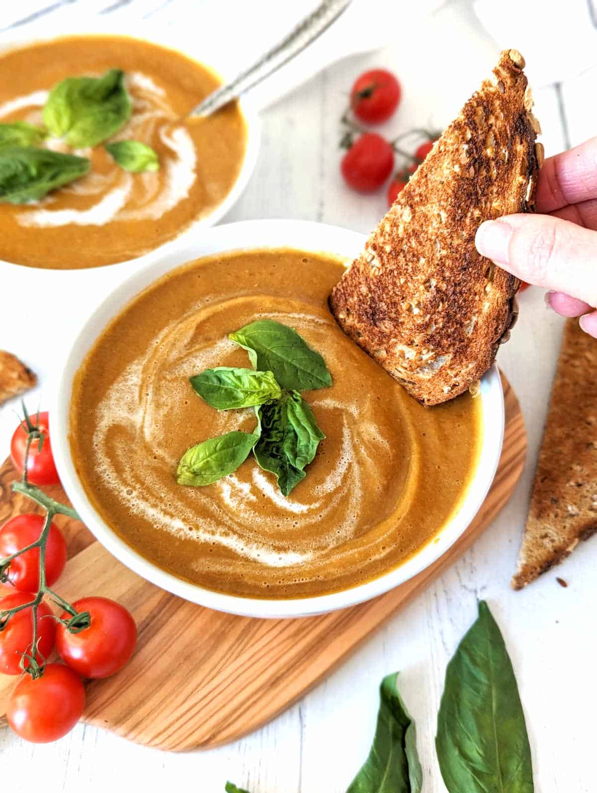 Roasted cherry tomato soup in a bowl with a swirl of coconut milk and topped with fresh basil leaves and a hand dipping a piece of toast into the bowl.