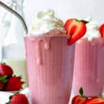 A glass full of strawberry banana milkshake topped with coconut whipped cream. Half of a strawberry placed on the rim of the glass.
