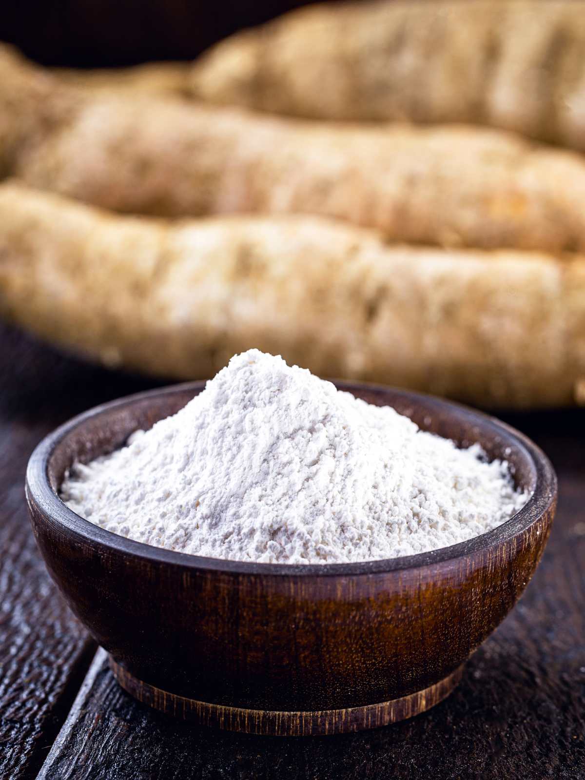 Tapioca flour in a wooden bowl with several cassava roots in the background.