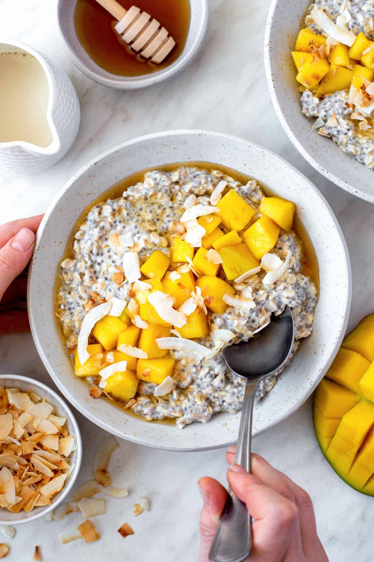 A hand dipping a silver spoon in a bowl of overnight oats with coconut milk.