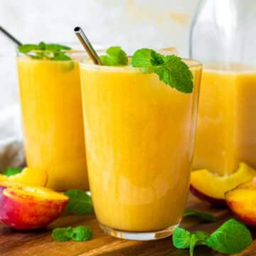 Two glasses filled with peach juice, topped with fresh mint leaves and served on a wooden board.