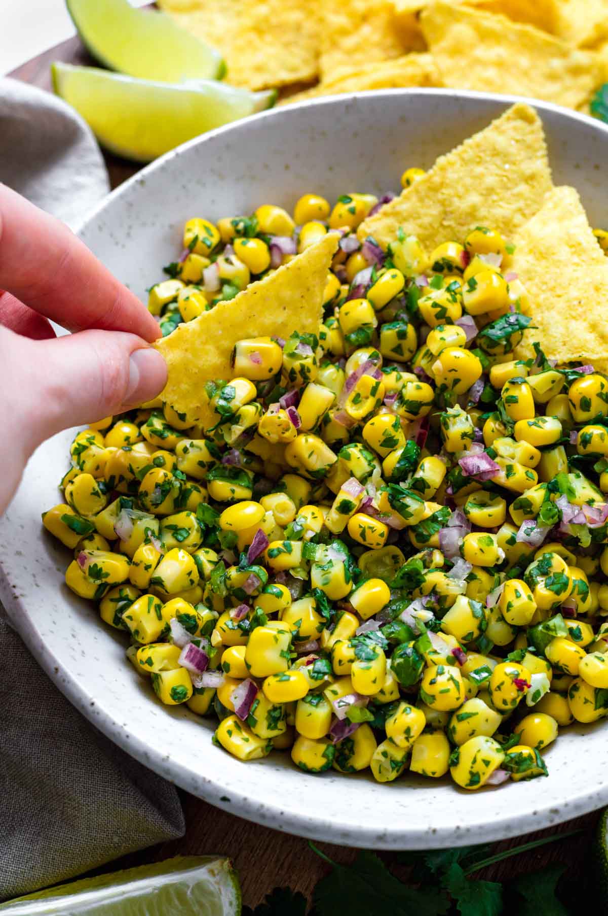 A hand holding a single tortilla chip and dipping it into a white bowl filled with corn salsa.