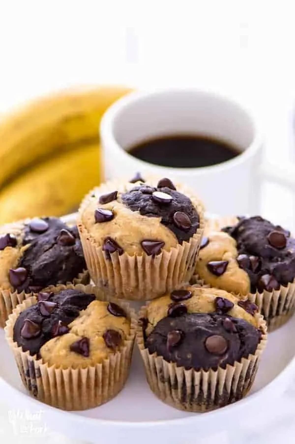 A plate of banana split muffins next to a cup of coffee and ripe bananas.