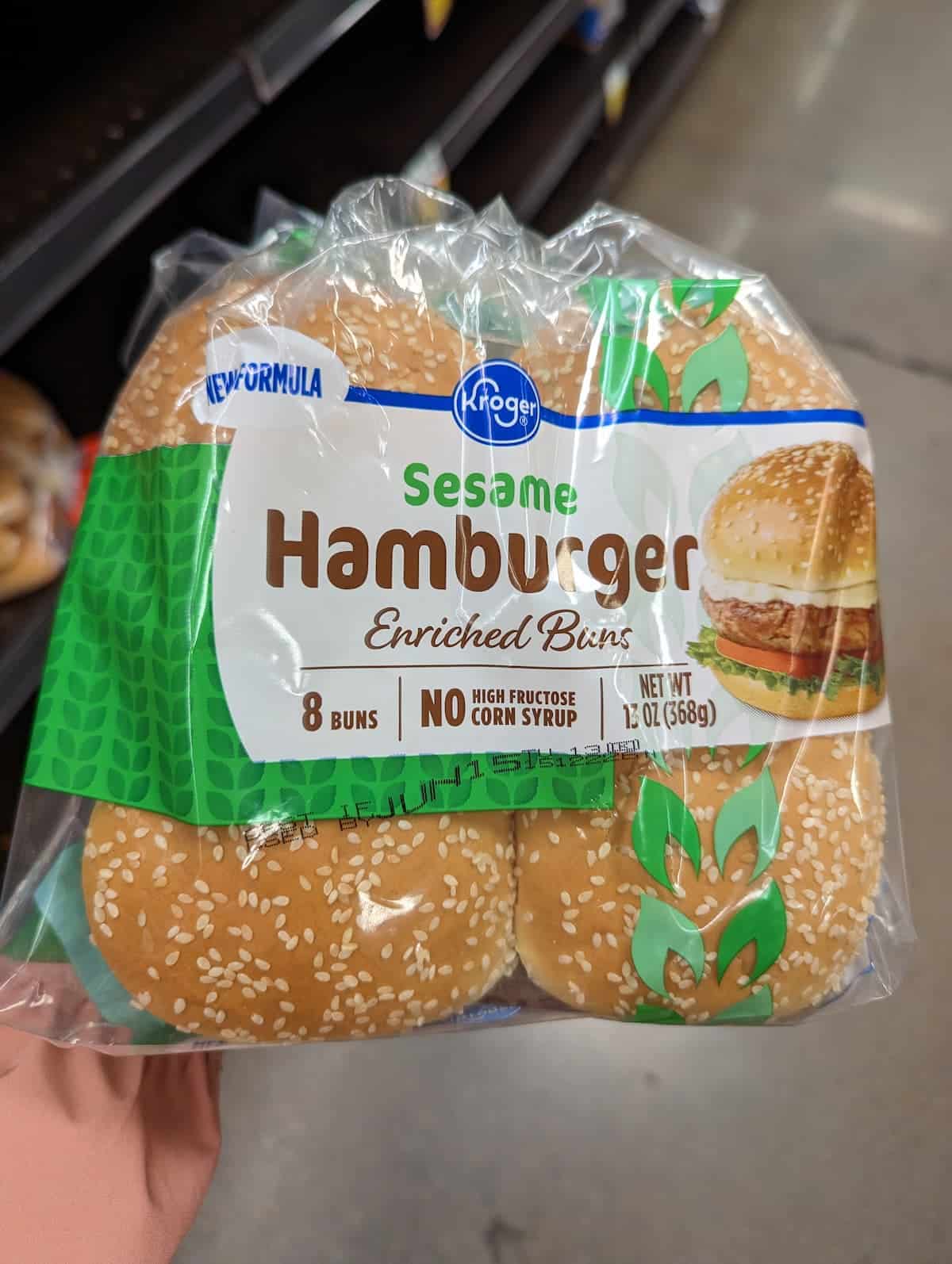 A hand holding a package of Kroger Sesame Hamburger Buns.